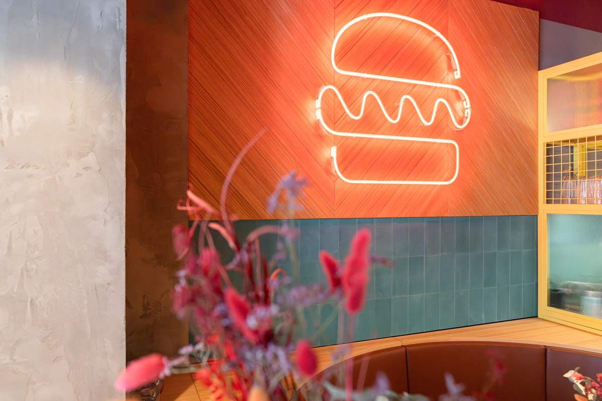 Im Vordergrund unscharf dekorative, bunte Trockenblumen, Im Hintergrund Neon-Burger-Symbol auf Holzpaneel und grüner Fliesenwand.