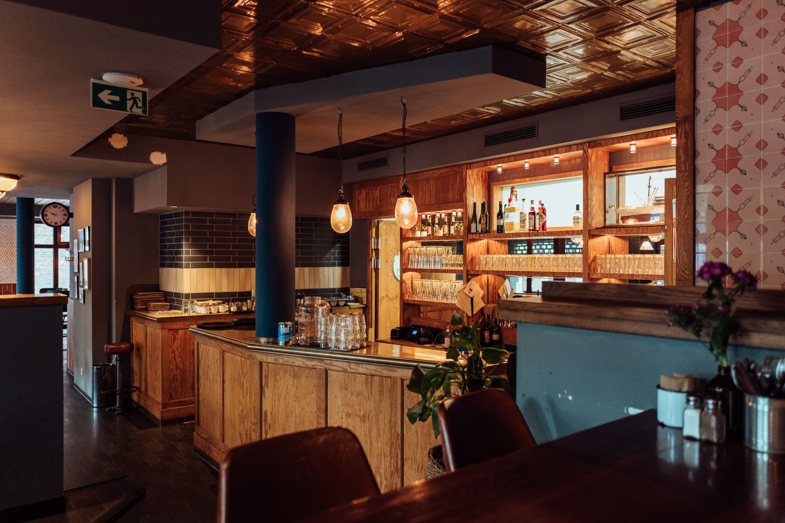 Barbereich von "Otto's Burger" mit Holztheke, offenen Regalen, Hängelampen, Fliesenwand und dekorativer Decke.