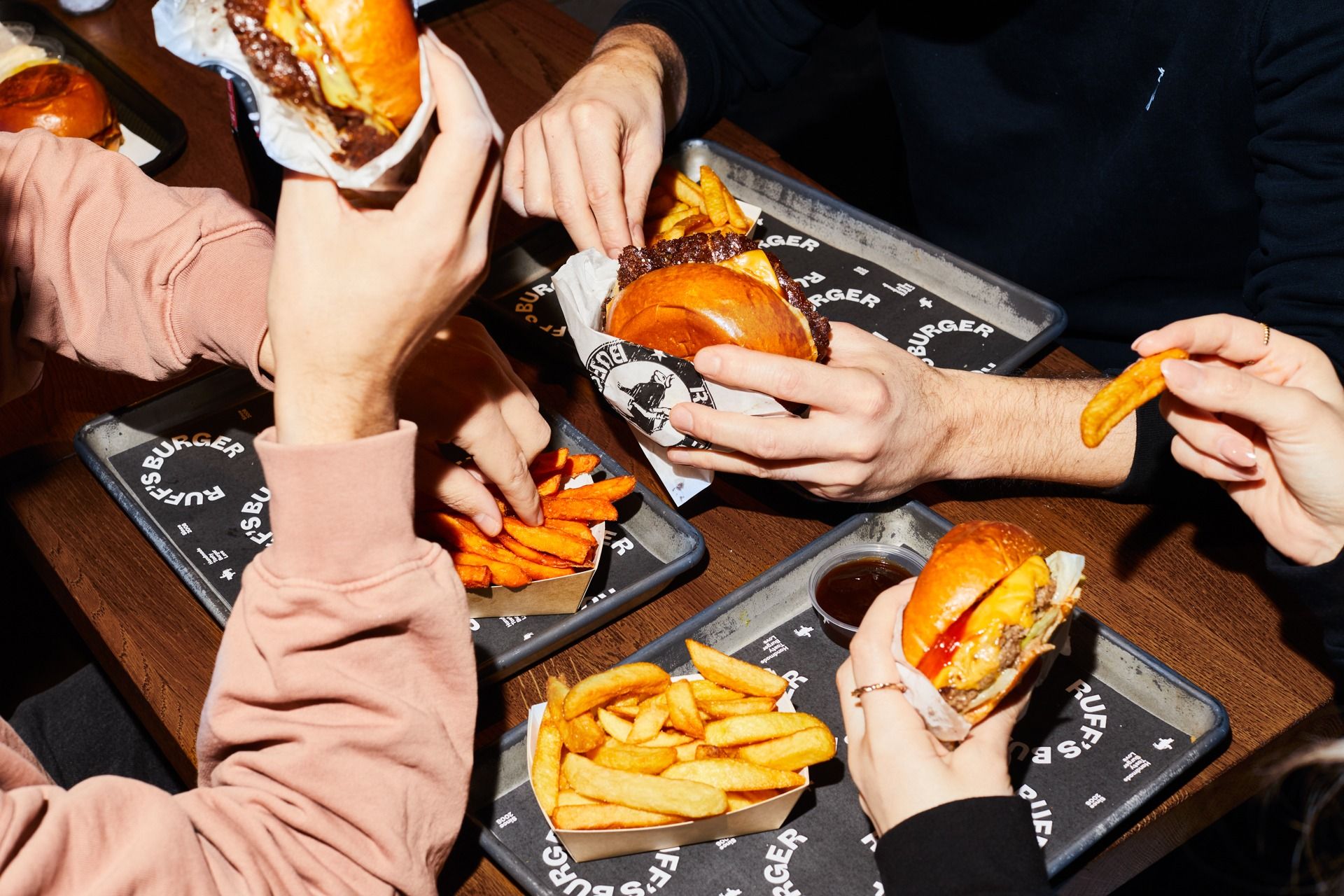 Drei Personen essen Burger und Pommes an einem Tisch, Tabletts mit "Ruff's Burger" Branding und Dipschalen.