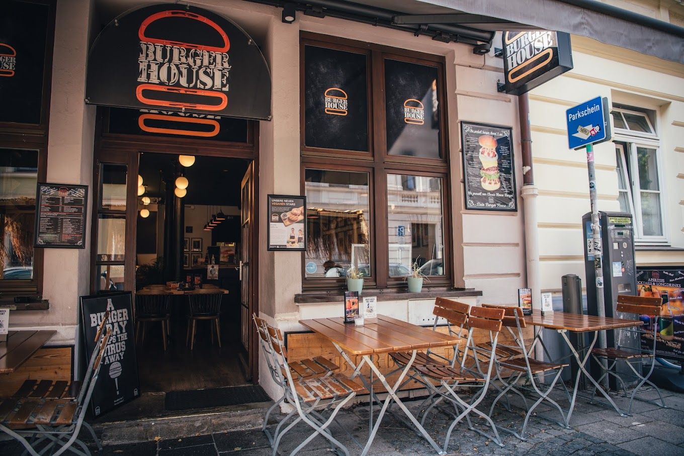 Außenansicht von "Burger House" mit Holztischen, Klappstühlen, beleuchtetem Logo und Speisekartentafeln am Eingang.