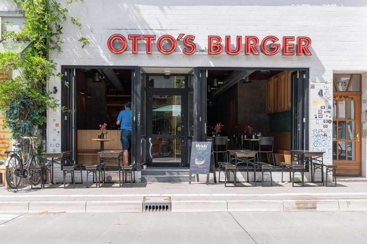 Außenansicht von "Otto's Burger" mit weißer Fassade, rotem Leuchtschriftzug, Außensitzbereich und Fensterfront mit Eingangstür.
