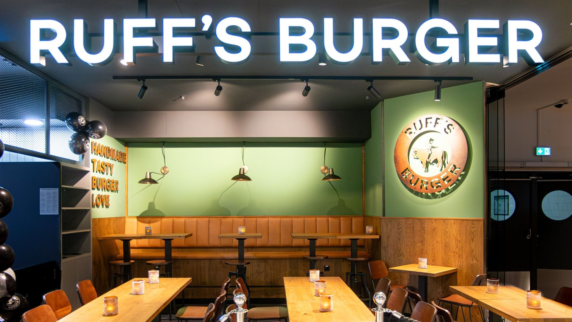 Innenansicht von "Ruff's Burger" mit Sitzbereich, grüner Wand, Wandlogo, Holztischen und leuchtendem Schriftzug.
