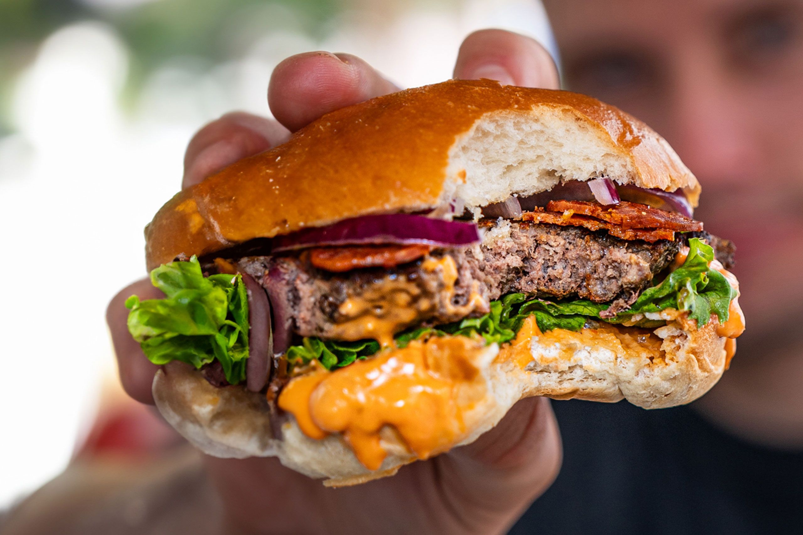 Nahaufnahme eines angebissenen Burgers mit Salat, Zwiebeln, Bacon, Patty und Sauce im Brioche-Bun.