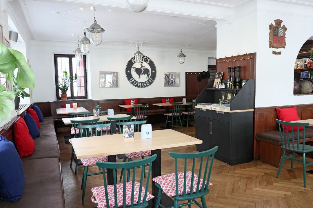 Innenansicht von "Ruff's Burger" mit Holztischen, grünen Stühlen mit roten Polsterkissen, Wandlogo und Parkettboden. Am Rand eine Servicestation mit Menükarten und Salz-, und Pfefferstreuern.