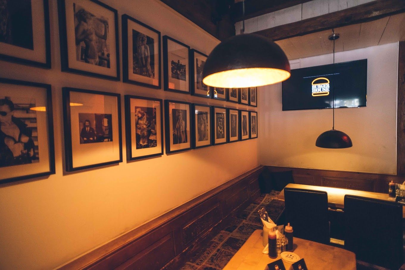 Innenbereich des Restaurants mit Holztisch, Wandgalerie aus Schwarz-Weiß-Fotos und TV-Bildschirm mit Logo.