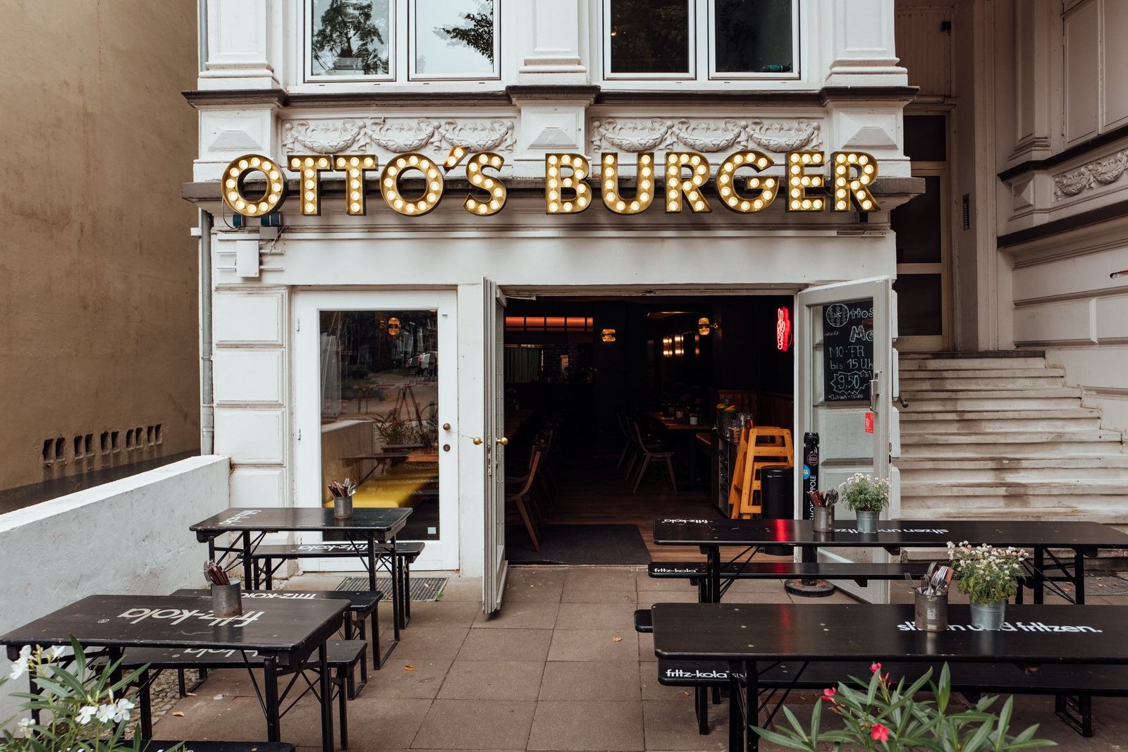Außenansicht von "Otto's Burger" mit beleuchtetem Schriftzug, Außensitzbereich mit schwarzen Holztischen und Stühlen.