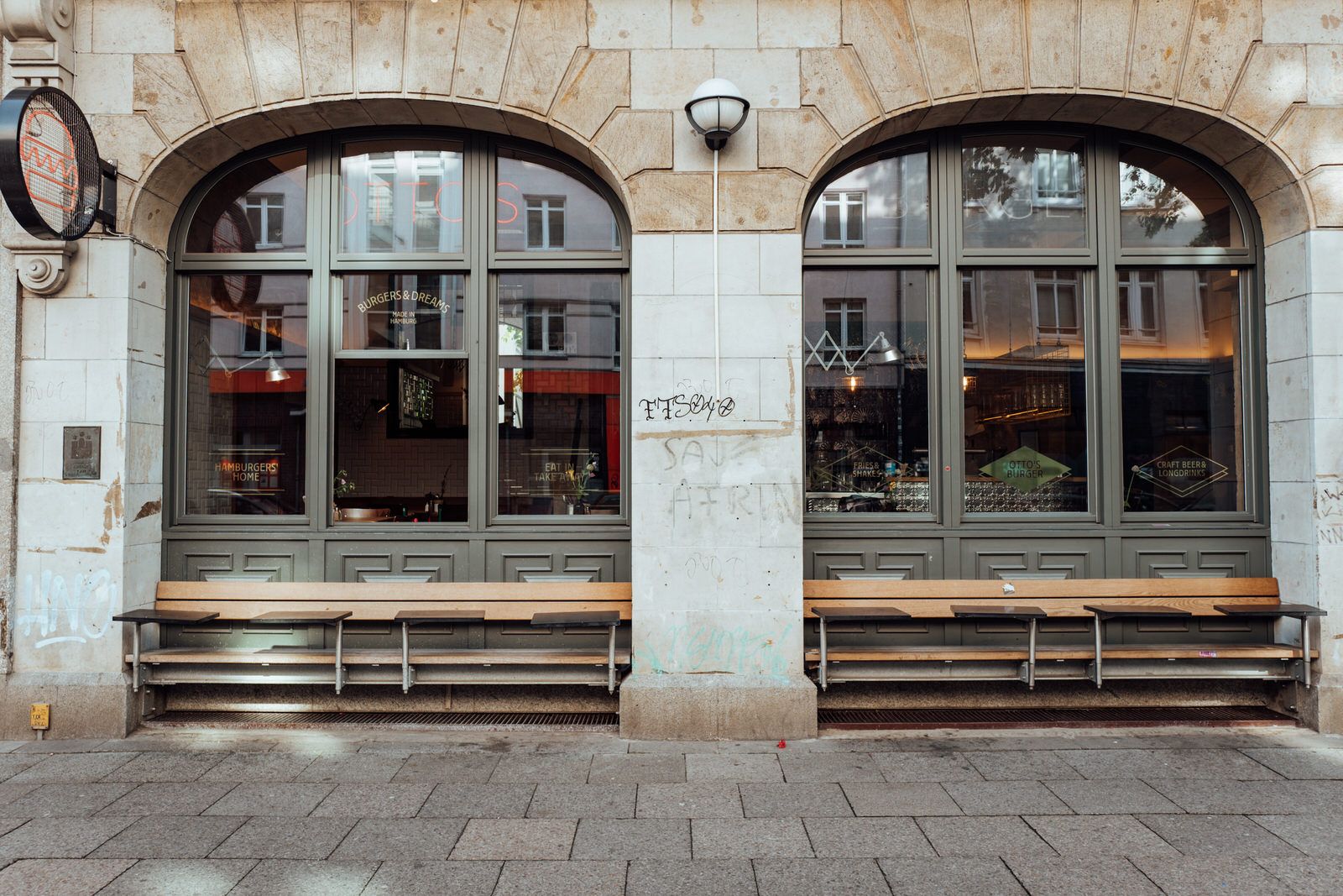 Frontale Außenansicht von "Otto's Burger" mit großer Fensterfront in Rundbögen, Sitzbänken und heller Natursteinfassade.