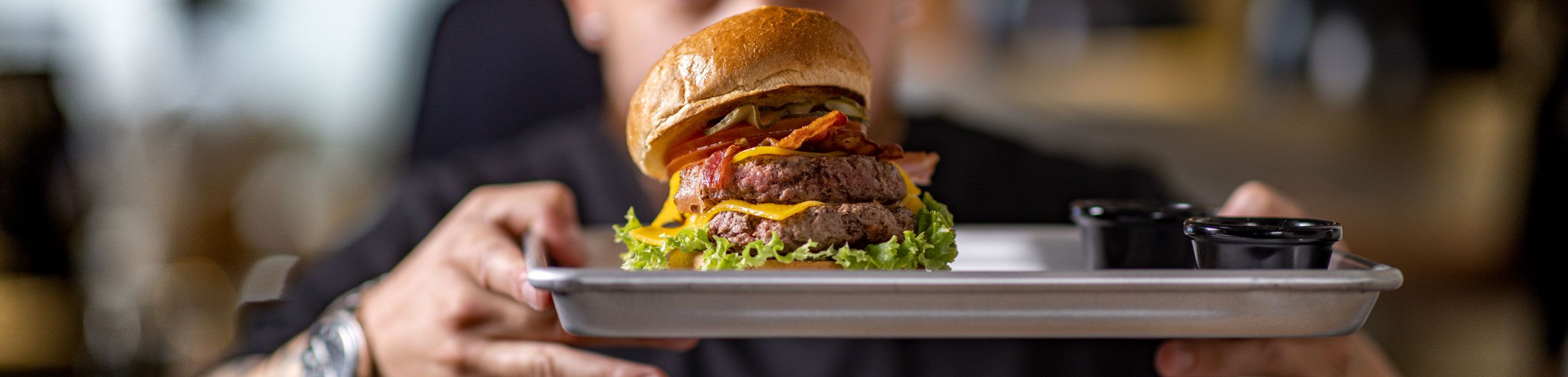 Burger auf Tablett mit Salat, Patty, Käse, Bacon, Gurken und Bun, gehalten von einer Person, im Hintergrund unscharf. Neben dem Burger stehen zwei schwarze Saucenschalen.