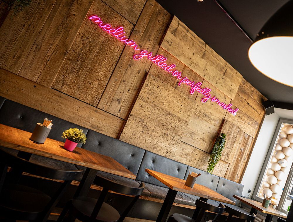 Innenansicht von "Ruff's Burger" mit Holzwand, schwarzer Polsterbank, Holztischen und pinkem Neon-Schriftzug "medium grilled or perfectly smashed".