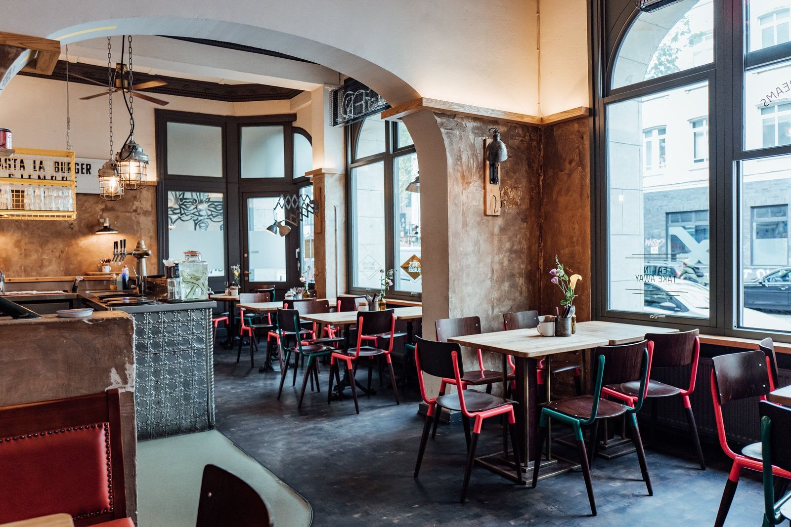 Innenansicht von "Otto's Burger" mit Holztischen, Fenstern, roten und grünen Stühlen, dunklem Boden und Thekenbereich im Hintergrund.