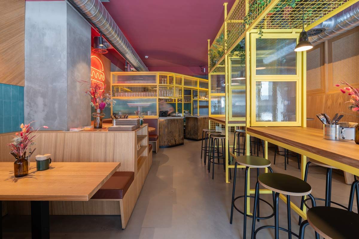 Innenansicht von "Otto's Burger" mit Holztischen, gelben Gittern, Barbereich, Neon-Elementen und offener Raumaufteilung.