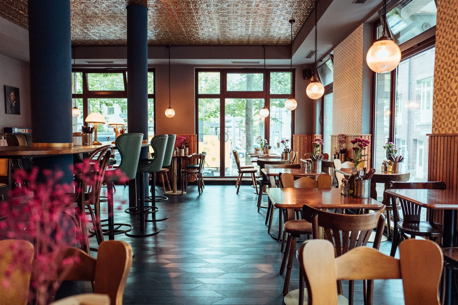 Innenansicht von "Otto's Burger" mit Holztischen, Stühlen, großen Fenstern, blauen Säulen und gemusterte Decke.
