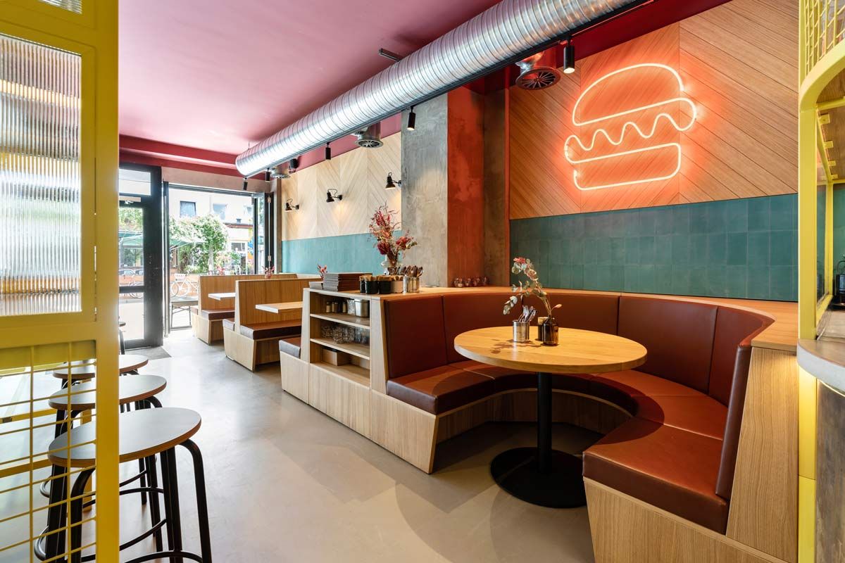 Innenansicht von "Otto's Burger" mit runder Sitzbank, Holztischen, Neon-Burger-Symbol, grünen Wandfliesen. Gelber Gitter-Hochtisch mit Barhockern.