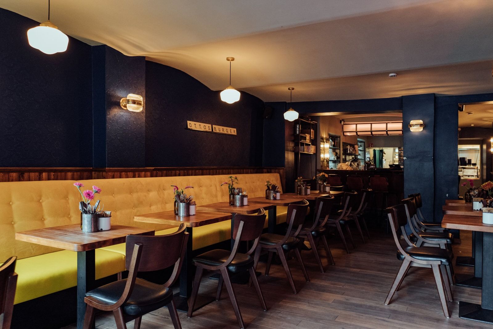 Innenansicht von "Otto's Burger" mit gelber Polsterbank, Holztischen, Holzstühlen und dunklen Wänden.