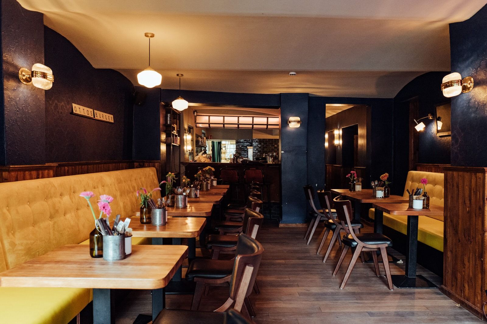 Eingangsbereich von "Otto's Burger" mit gelber Polsterbank, Holztischen, Holzstühlen, Dekoration und Blick Richtung Barbereich.