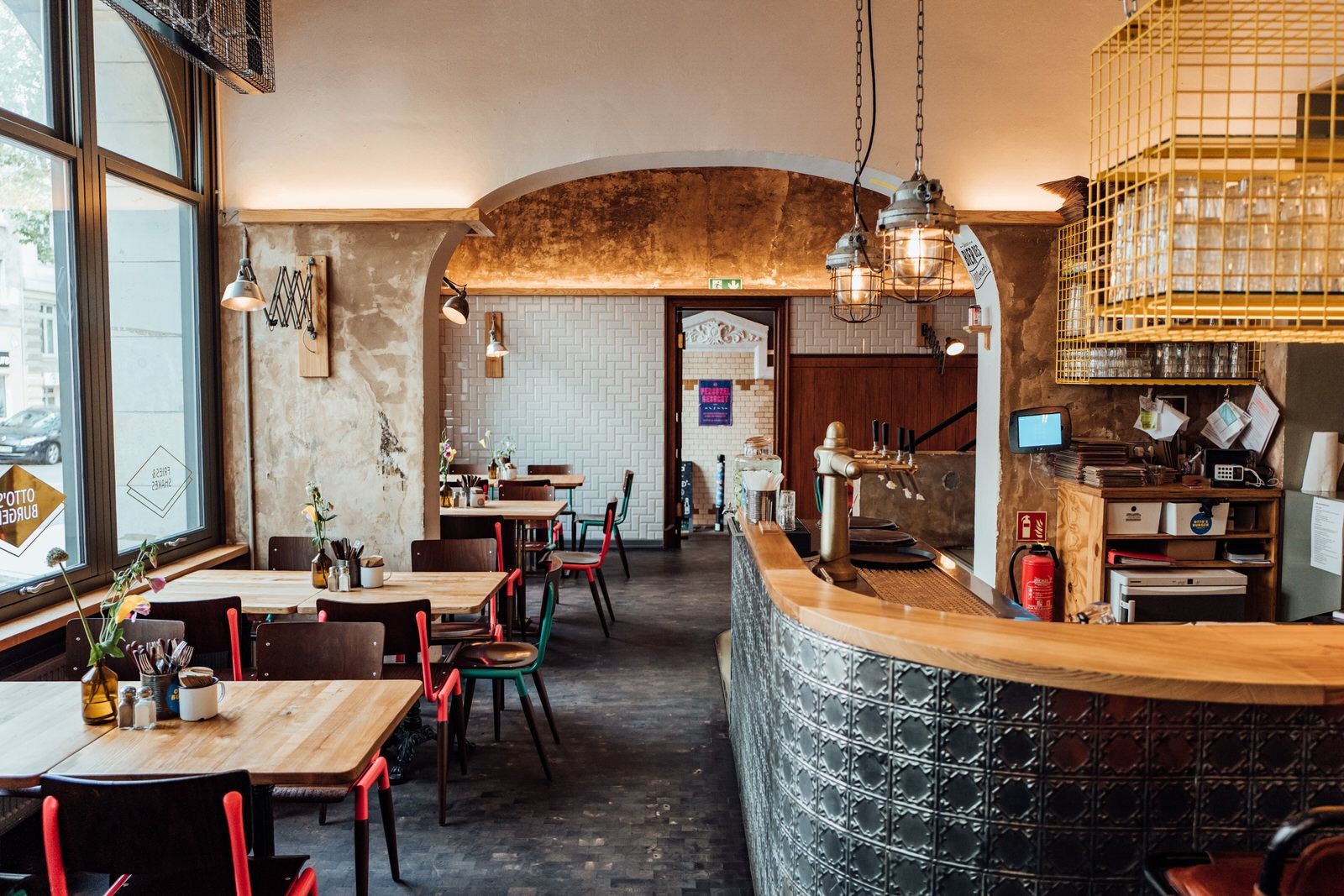 Innenansicht von "Otto's Burger" mit Holztheke, Holztischen, roten und grünen Stühlen, Fenstern, Metalllampen, Gläserregal über der Theke.